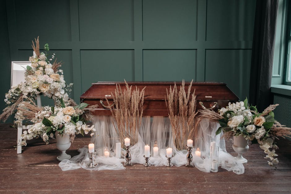 A floral arrangement placed on a funeral setting featuring two large white vases filled with white roses, lilies, and greenery, accented with beige pampas grass. The flowers are arranged with some spi