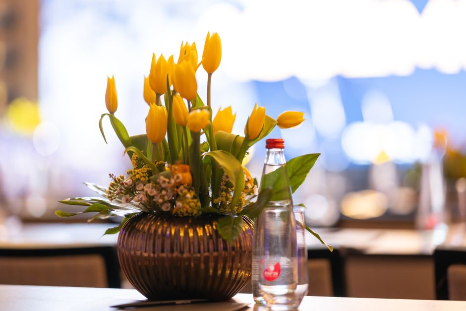 A floral arrangement featuring bright yellow tulips with closed buds and elongated petals, set in a ribbed, dark amber glass vase. The bouquet is complemented by small clusters of pink and yellow fill