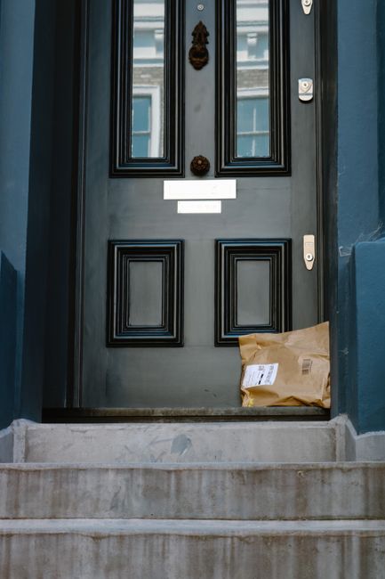 A brown paper package with a shipping label rests on a stone doorstep in front of a black wooden door with decorative paneling and a brass door knocker, part of a residential entrance. The door is fra