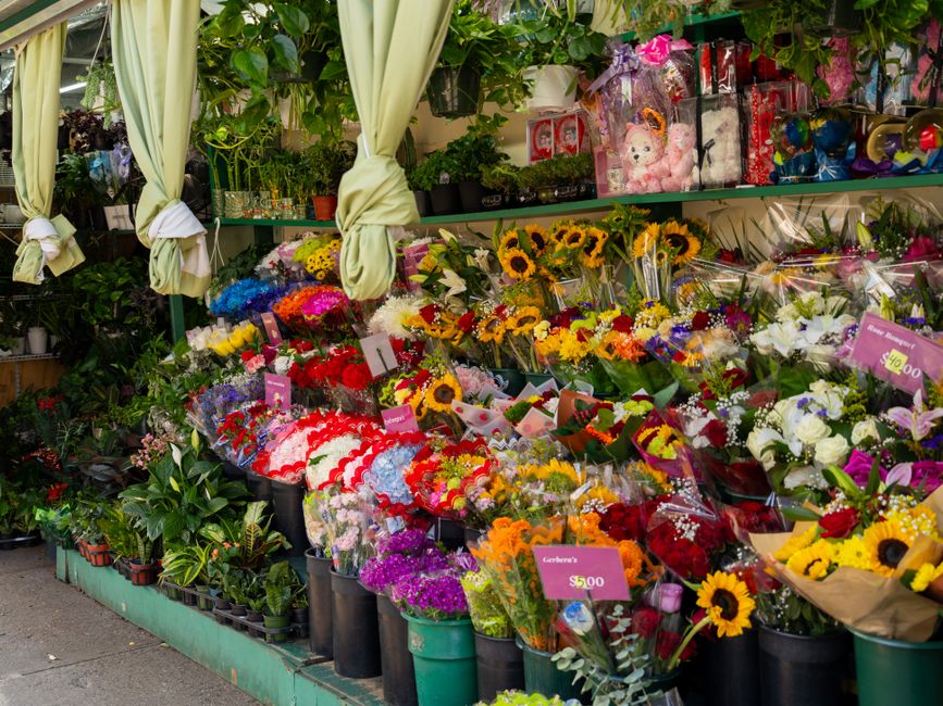A vibrant outdoor display of fresh flower bouquets arranged on a shop stand, featuring a variety of colorful blooms including bright yellow sunflowers, red and pink roses, white lilies, purple and pin