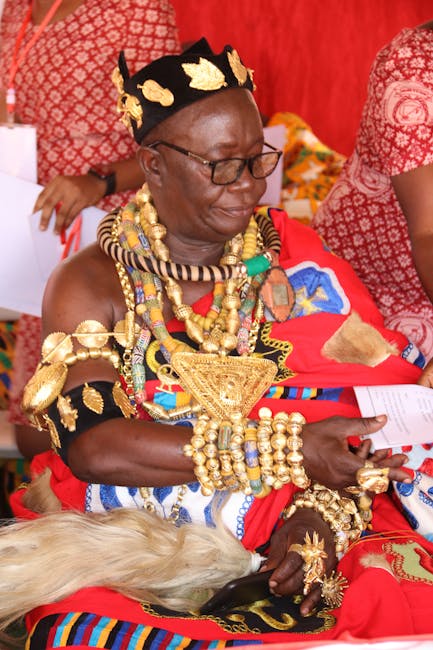 A woman wearing traditional Maasai attire with a vibrant red shuka, adorned with elaborate gold jewelry including necklaces, bracelets, and a headpiece, sitting outdoors against a red fabric backdrop.