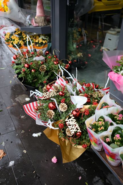 A festive arrangement of fresh flowers and holiday decorations displayed outside a flower shop, featuring a bouquet with red berries, pinecones, and greenery wrapped in gold paper with a striped red a